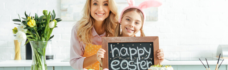 panoramic shot of cheerful mother near cute daughter holding chalkboard with happy easter lettering