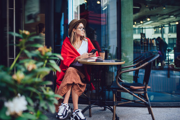 Insightful hipster woman in hat texting smartphone while sitting at cold cafe terrace