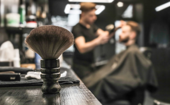 Soft Brush. Close Up Photo Of A Big Soft Shaving Brush Standing On A Barber’s Workplace While A Hairstylist Is Grooming His Client’s Beard.