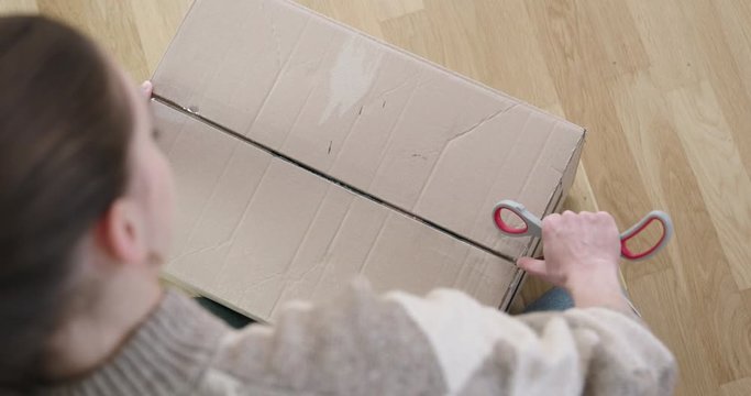 Young woman cuts postal parcel open with scissors and opens the cardboard box. Cardboard package contains plastic air bags. Over the shoulder, static shot.