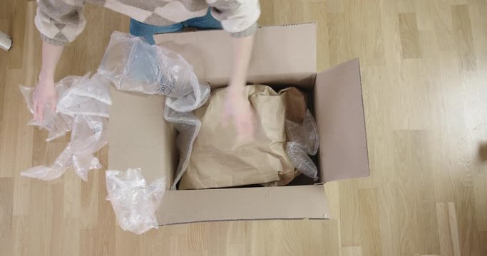 Female person opening package and taking out protective material (air bags). Woman grabs package and holds it up. Static, top down shot.
