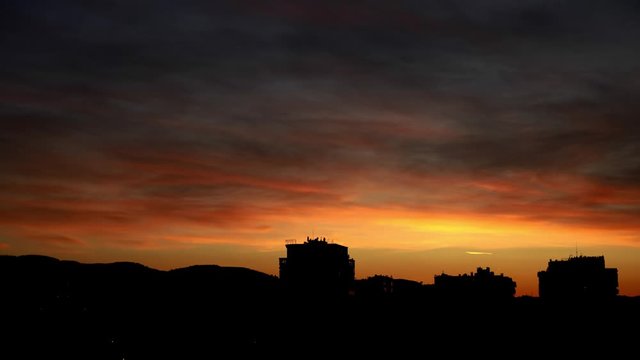 Silhouette of city buildings at dramatic sunset with glowing yellow red sky under dark grey clouds moving fast
