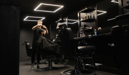 Black stylish room. Side view photo of a young professional barber trimming hair of his male...