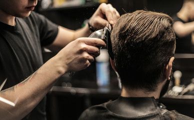 Creative process. Side view photo of a young barber trimming his customer’s hair with an electric shaver and a comb.
