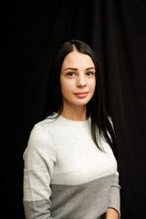 Portrait of serious beautiful female with black hair, has minimal makeup, looks calmly at camera, wears white jumper, stands against black background, being deep in thoughts