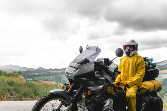 Motorcyclist traveler girl wearing yellow raincoat and sitting in a motorcycle saddle. Motorbike on mountain road. Extreme travel tour. Biker equipment. Dramatic cloud, copy space
