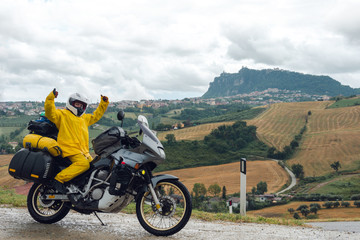 Happy motorcyclist traveler girl wearing yellow raincoat and sitting in a motorcycle saddle. Destination scene. Extreme travel tour. Biker equipment. View of the mountain. San Marino, Italy.