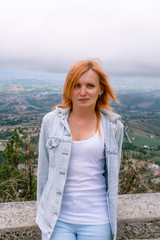 Vertical photo. Portrait of a blonde girl. Dressed in a light denim jacket, blue pants and a white tank top. Posing on top of a mountain overlooking the city of San Marino, Italy