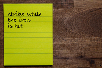 yellow sticker with lines on a dark wooden background with the handwriting of the wisdom of life: strike while the iron is hot