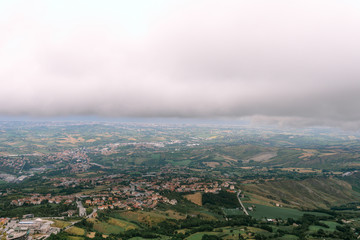 View from far away to the city, a cloud over the city, small houses of fields and forests. Modern San Marino Suburban districts and Italian hills view from above. Horizontal shot