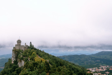 Fototapeta na wymiar View from the top of the mountain to the medieval fortification. Dramatic mystical weather with fog. Castle in San Marino (Seconda Torre) Second Tower, Copy space