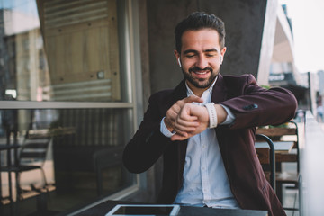 Cheerful caucasian bearded businessman sitting on cafe terrace using modern digital devices for checking mails and notification,smiling young man in formal wear looking at his wristwatch display.