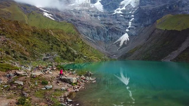 Aerial, drone shot following a woman hiking on the rocky shore of lake Humantay, overcast day, in Andes,  Cusco region, Peru