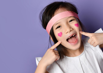 Portrait of happy smiling frolic asian Korean kid girl laughing pointing fingers on cheeks with red hearts sign on cheek
