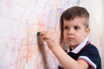 The child draws on the wall with a crayon. The boy is engaged in creativity at home