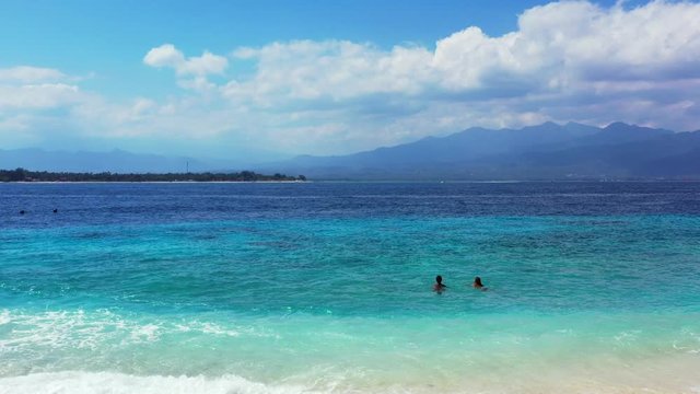 Girls swim on shallow turquoise lagoon near shore of tropical island under bright sky with white clouds hanging over mountains in Bali