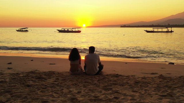Young couple in love sitting on tranquil exotic beach, watching beautiful sunset with orange sky reflecting on calm sea