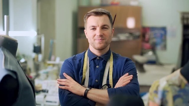 male clothing designer in sewing shop folds his hands and looks at the camera with a smile