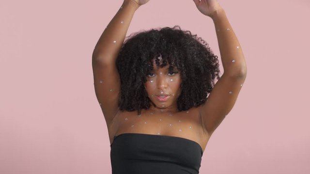 Mixed race black woman with curly hair covered by crystal makeup on pink background in studio Dancing in slow motion from 60 fps. Fashion makeup