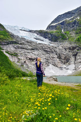 The young girl looks at the Boyabreen glacier, which is the sleeve of large Jostedalsbreen glacier and wild yellow flowers in the foreground. Melting glacier forms the lake with clear water. Norway.