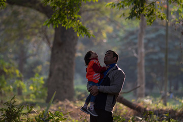 Naklejka na ściany i meble An Indian brunette father and his baby boy in winter garments enjoying themselves in winter afternoon in light and shadow in green forest background. Indian lifestyle and parenthood.