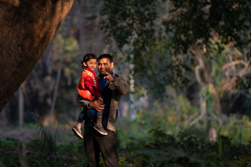 An Indian brunette father and his baby boy in winter garments enjoying themselves in winter afternoon in light and shadow in green forest background. Indian lifestyle and parenthood.