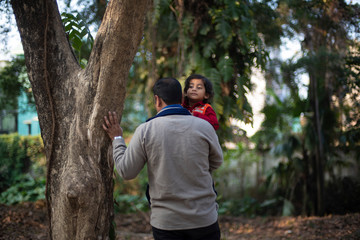 Back side of Indian brunette father taking his baby boy in lap in winter garments in winter afternoon on a dry grass field in green forest background. Indian lifestyle and parenthood.