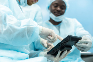 A team of doctors in uniform and medical masks looks at a surgical plan on an electronic tablet