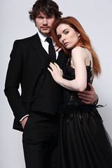 Sexy bright foxy hairstyle female woman in fashion black long skirt dress hugging her handsome man in black suit clothing on light blue studio background. Closeup