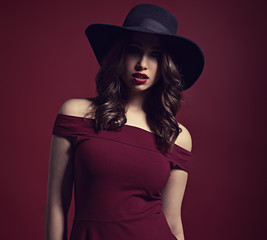 Fototapeta na wymiar Beautiful makeup woman with long brown curly volume hair style, red lipstick, and black smokey eyes in red dress and blue elegant hat. Closeup portrait on dark