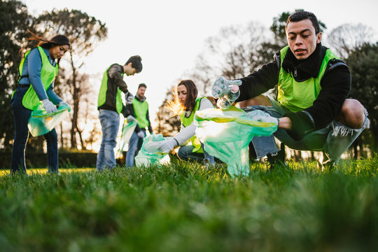 Group of friends during a volunteer garbage collection event in a park at sunset - Millennial having fun together - Happy people cleaning area with bags - Ecology concept