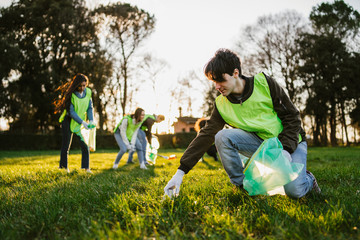 Group of friends during a volunteer garbage collection event in a park at sunset - Millennial having fun together - Happy people cleaning area with bags - Ecology concept - 323942213