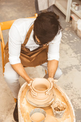 A young male potter is engaged in craft in his workshop on a potter's wheel and makes a clay product