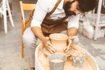 A young male potter is engaged in craft in his workshop on a potter's wheel and makes a clay product