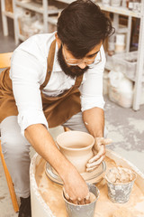Obraz na płótnie Canvas A young male potter is engaged in craft in his workshop on a potter's wheel and makes a clay product