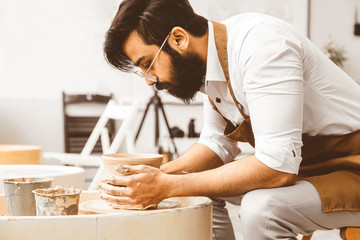 A young male potter is engaged in craft in his workshop on a potter's wheel and makes a clay product
