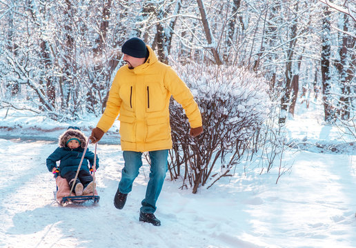 Winter family walk outdoors: photo of dad running along snowy road and pulling sled with toddler child sitting in it. Father and son sledding and having fun together. Focus on child face. Copy space