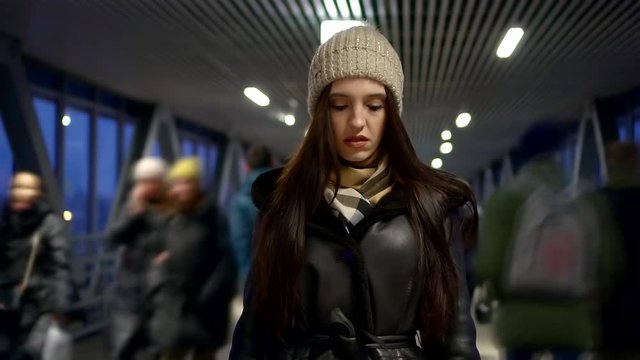 A close-up portrait of a young dark-haired girl standing on a bridge that is an elevated pedestrian crossing. She's wearing a hat and a black leather coat. People pass by it. Accelerated shooting.