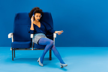 african american sitting on seat, listening to music and holding smartphone on blue background