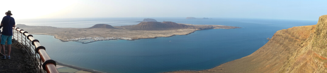 Fototapeta na wymiar A panoramic view of the volcanic island of La Graciosa meaning graceful in Spanish off Lanzarote,Spain.A male tourist wearing a hat stands by a railing on a cliff edge admiring the scenery.Image