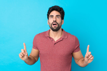 Young handsome man with beard over isolated blue background surprised and pointing up