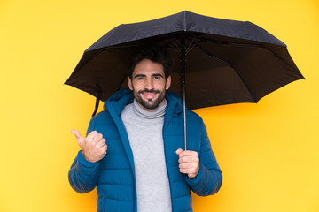 Man holding an umbrella over isolated yellow background pointing to the side to present a product