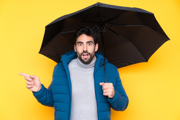 Man holding an umbrella over isolated yellow background surprised and pointing finger to the side