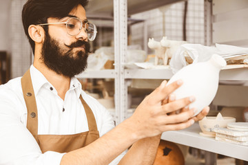 A young attractive male potter with a beard and mustache works in his workshop. Holds and inspects...