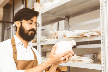 A young attractive male potter with a beard and mustache works in his workshop. Holds and inspects...