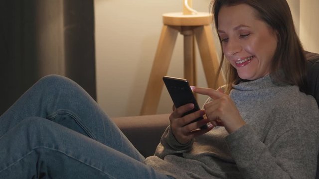 Smiling woman lying on the couch in a cozy room and using smartphone for surfing internet in the evening. Relaxation and lifestyle technology.