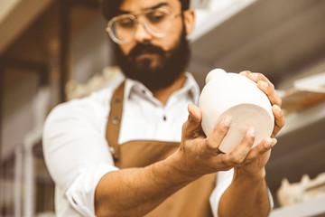 A young attractive male potter with a beard and mustache works in his workshop. Holds and inspects...