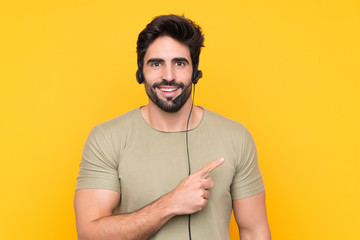Telemarketer man working with a headset over isolated yellow background pointing finger to the side