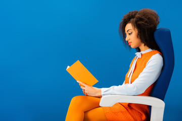 side view of african american in retro dress sitting on seat and reading book isolated on blue