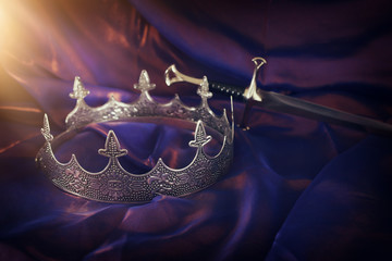 low key image of beautiful queen/king crown and sword over dark royal purple delicate silk. fantasy...